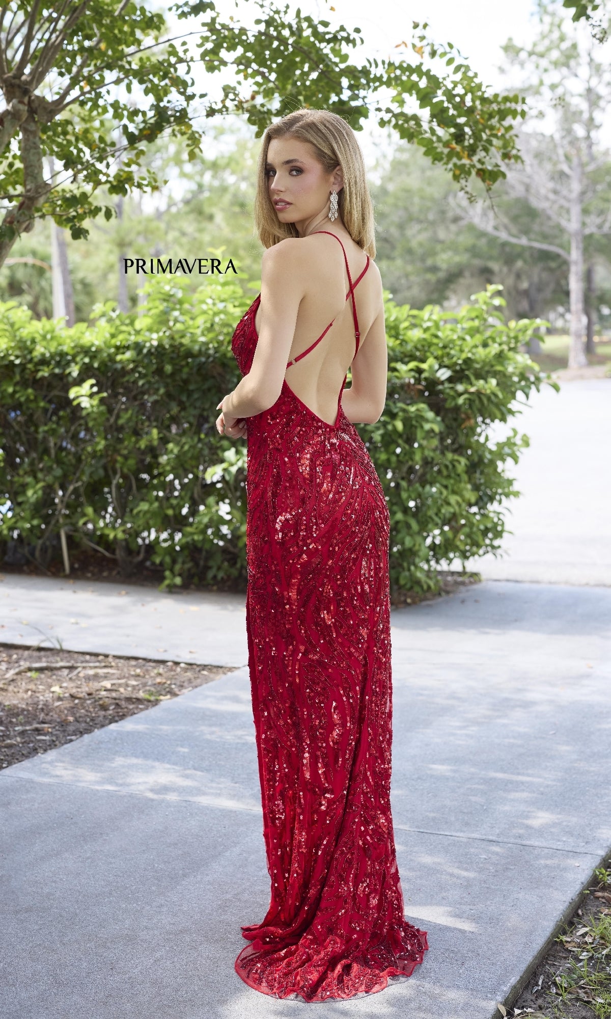 A woman wearing the Long Prom Dress: Primavera 4371, a red sequin gown with an open back, stands outdoors on a paved path beside green bushes and trees.